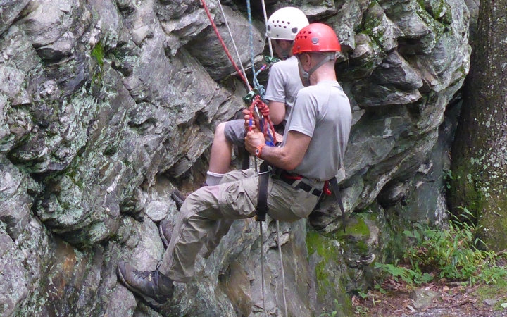 Two people wearing safety gear are secured by ropes as they sit back into their harnesses while taking a break from rock climbing.